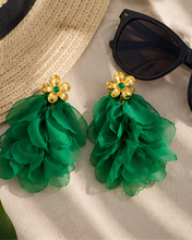 Load image into Gallery viewer, A pair of green ruffled flower drop earrings with gold flower-shaped tops and small green center stones, styled on a beige fabric surface with a straw hat and black sunglasses nearby.