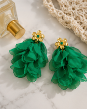 Load image into Gallery viewer, A pair of green ruffled flower drop earrings with gold flower-shaped studs and small green gem accents, displayed on a white marble surface with a perfume bottle and crochet bag as props.