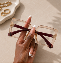 Load image into Gallery viewer, A hand holding a pair of oversized square sunglasses with a translucent blush tan frame and dark burgundy temples, set against a neutral beige background with gold jewelry props.
